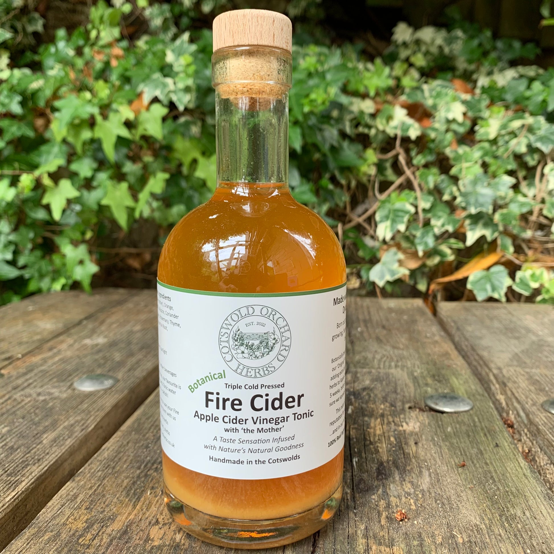 Bottle of Botanical Fire Cider on a wooden surface with greenery in the background