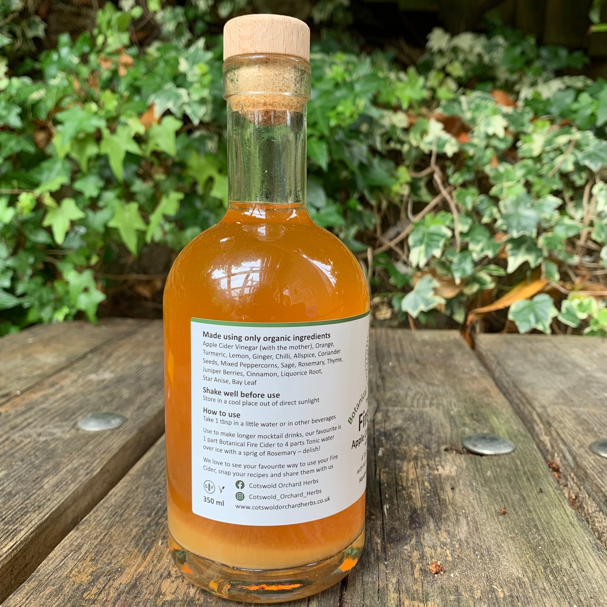 Bottle of organic Botanical Fire Cider vinegar with a cork lid on a wooden surface with greenery in the background