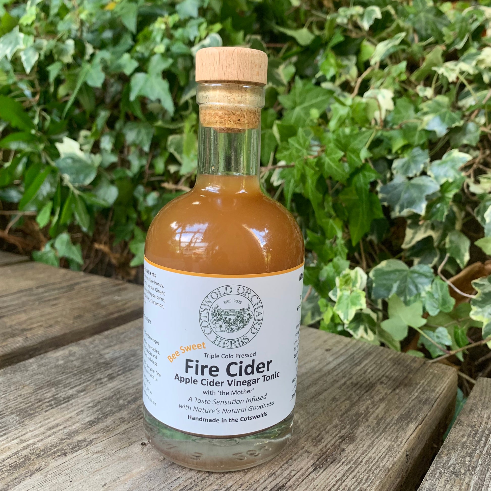 Bee Sweet Fire Cider 250ml – organic herbal tonic in clear glass bottle with cork top, photographed on rustic wooden surface with lush green ivy in background