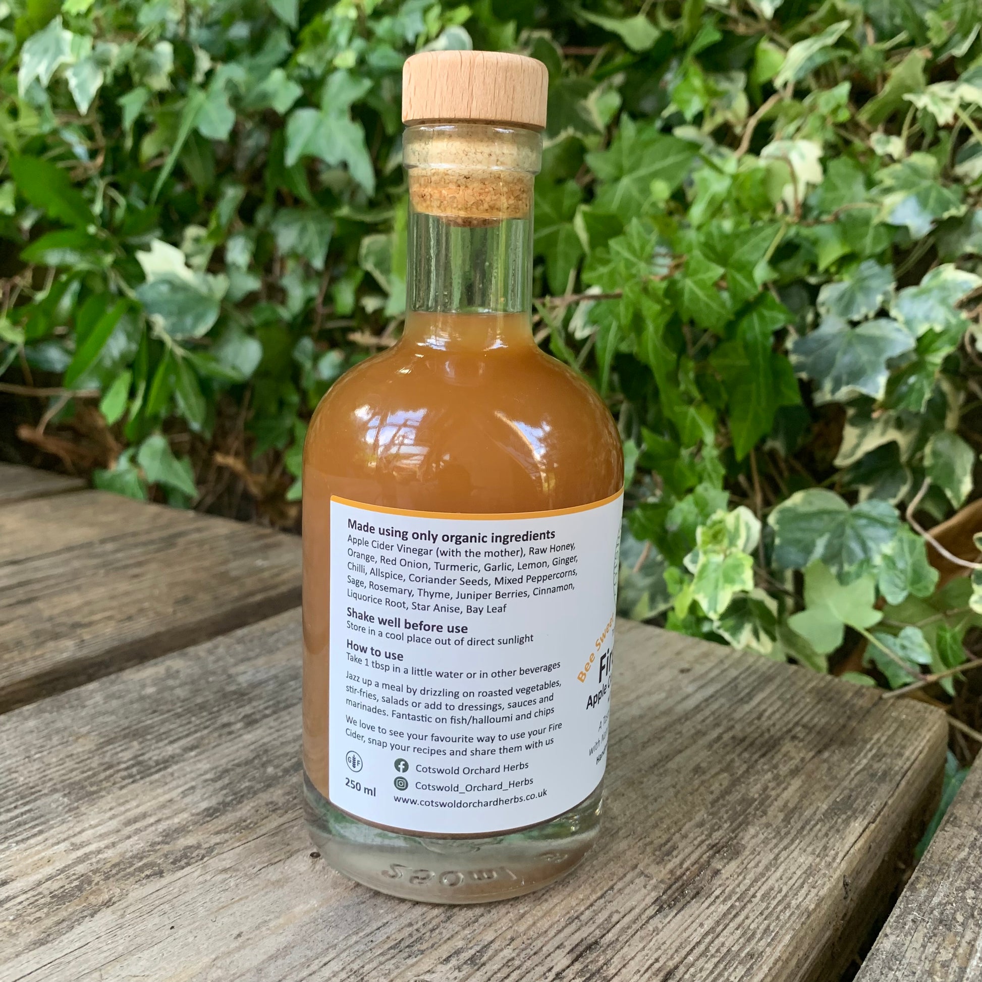 Back label of Bee Sweet Fire Cider 250ml – ingredients and usage instructions visible on bottle, positioned outdoors on wooden bench with green foliage