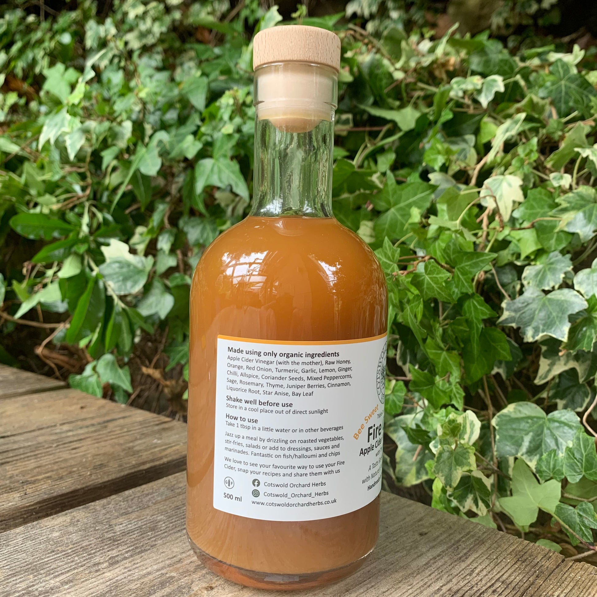 Side view of Bee Sweet Fire Cider 500ml bottle – label listing organic ingredients, suggested use, and storage info, positioned on rustic wooden bench with garden ivy background