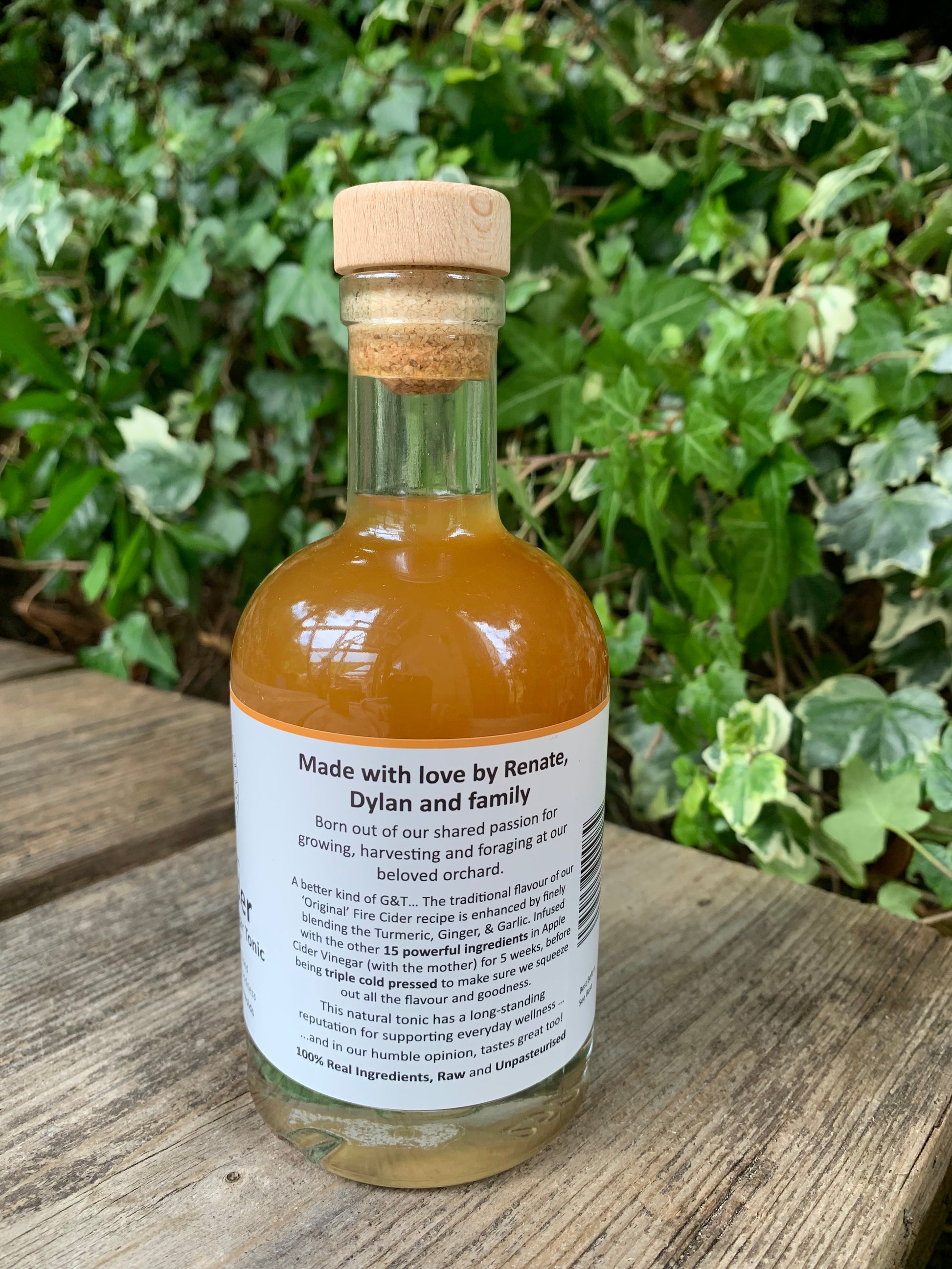 Back label of G&T Boost Fire Cider 250ml – handcrafted tonic made with apple cider vinegar, turmeric, garlic, and ginger, created by Renate, Dylan and family, set on a wooden bench with leafy background
