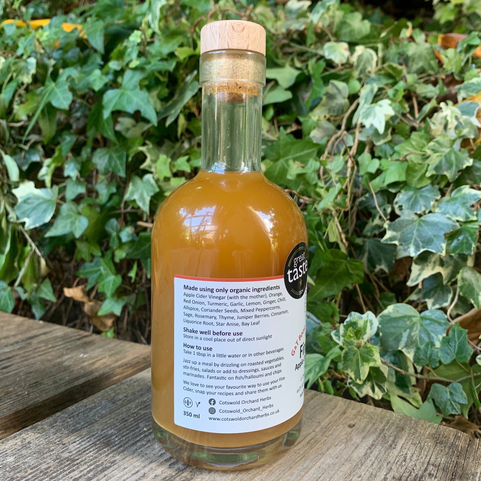 Side label of G&T Boost Fire Cider 350ml bottle – listing organic ingredients and suggested uses, with Great Taste award badge, set on rustic wooden bench outdoors