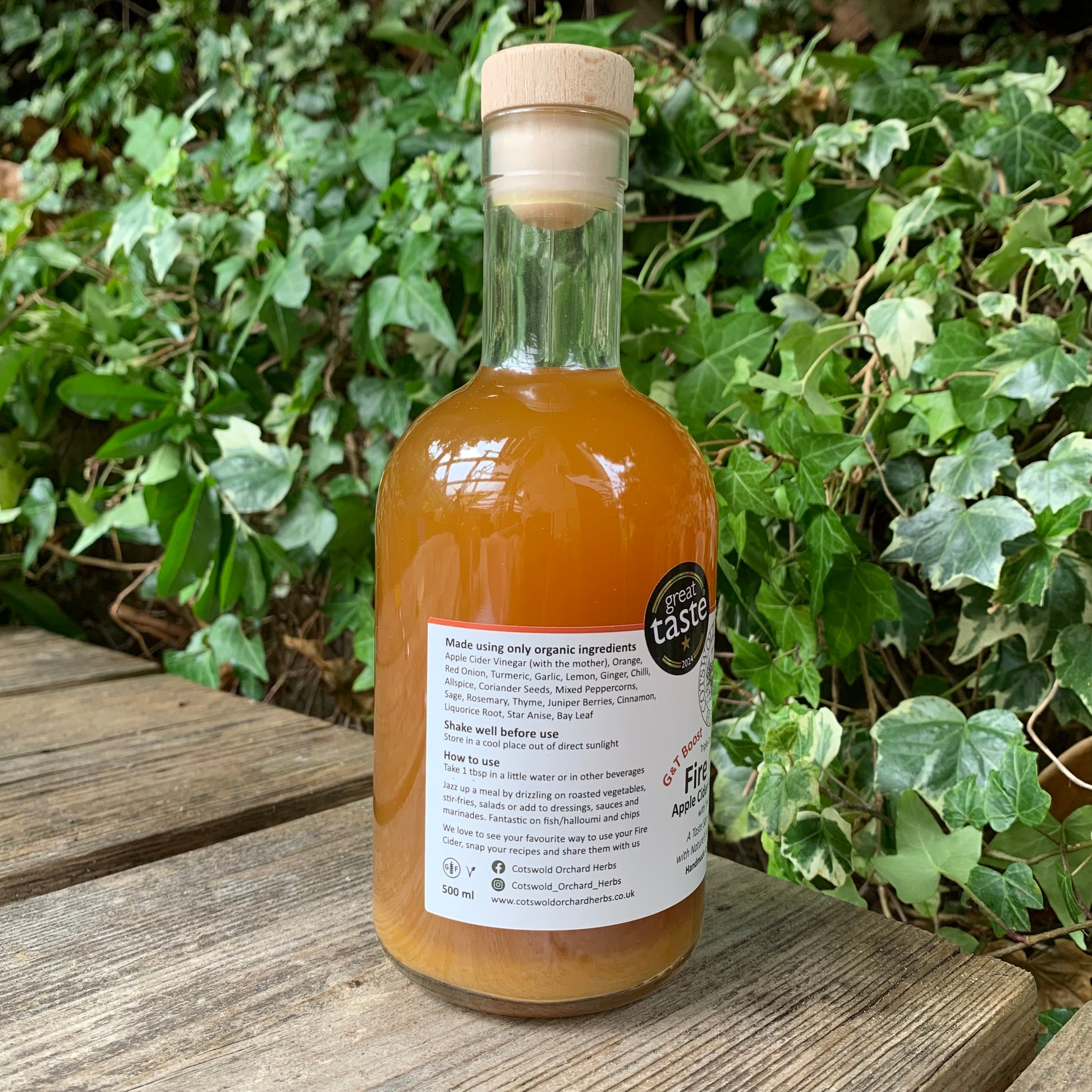 Side view of G&T Boost Fire Cider 500ml – label showing organic ingredients and usage instructions, with Great Taste award seal, photographed on rustic wooden bench in natural garden setting