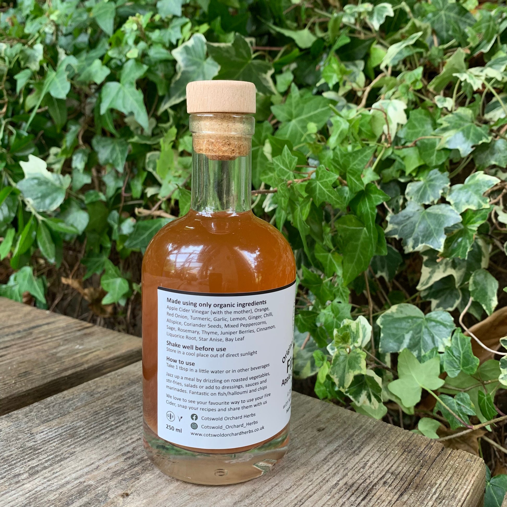 Side label of Original Fire Cider 250ml bottle – listing organic ingredients and usage suggestions, placed outdoors on rustic wood with green garden foliage