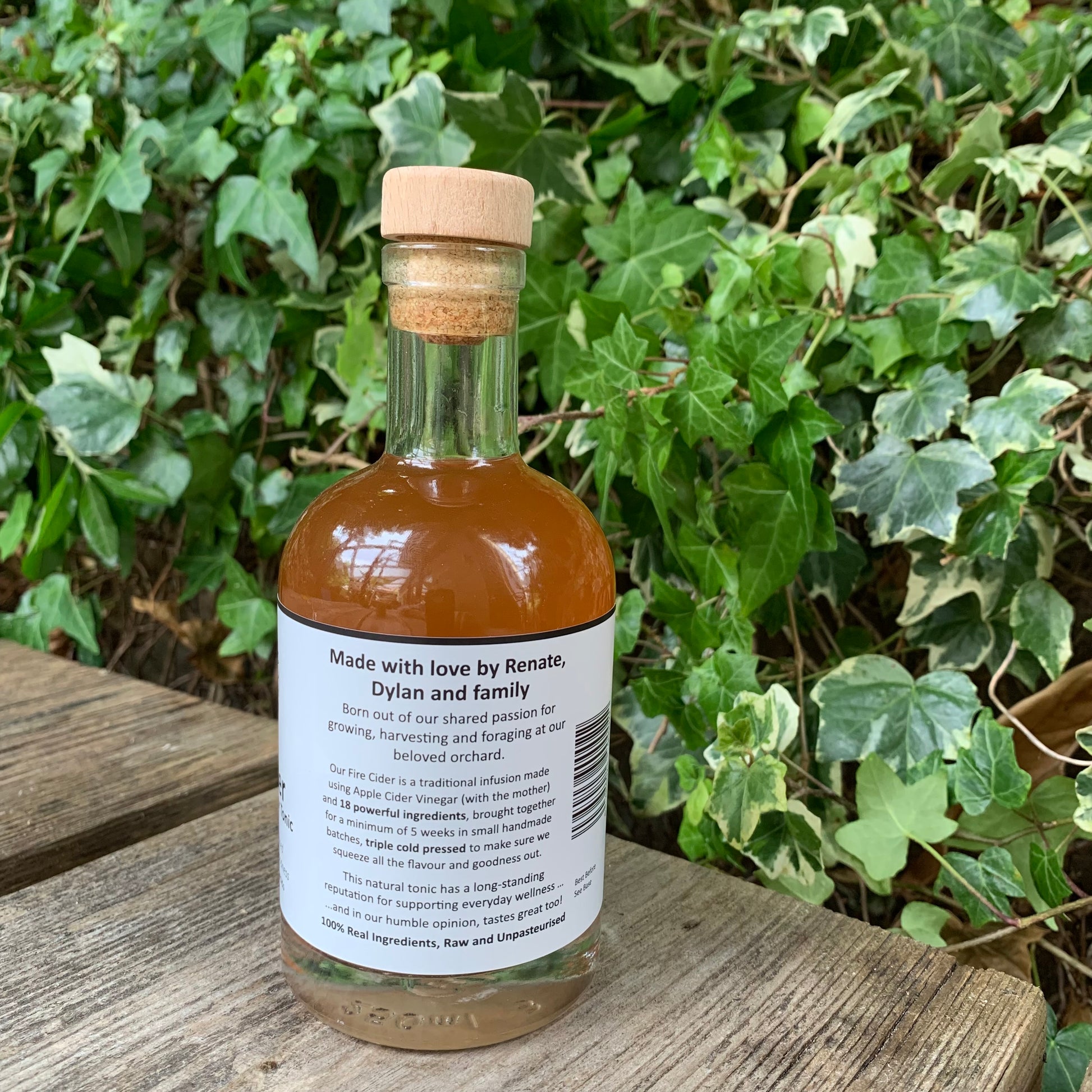 Back label of Original Fire Cider 250ml – small-batch apple cider vinegar tonic handcrafted by Renate, Dylan and family, displayed on wooden bench with natural ivy backdrop