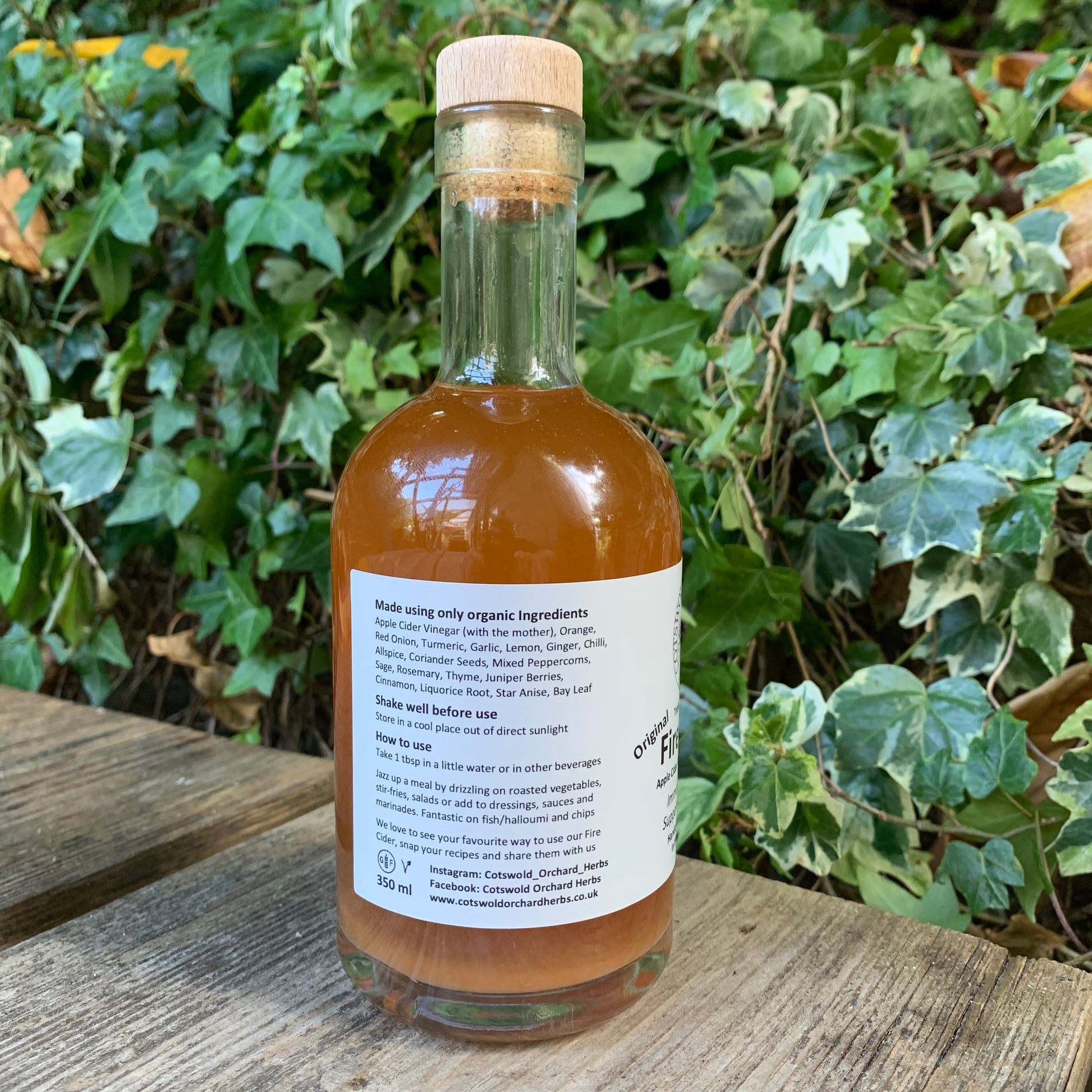 Side label of Original Fire Cider 350ml bottle – shows list of organic ingredients and suggested usage, placed on wooden surface with leafy garden background