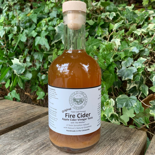 Front-facing 500ml bottle of Original Fire Cider – apple cider vinegar tonic infused with natural herbs, handcrafted by Cotswold Orchard Herbs, displayed on rustic wooden bench with ivy background