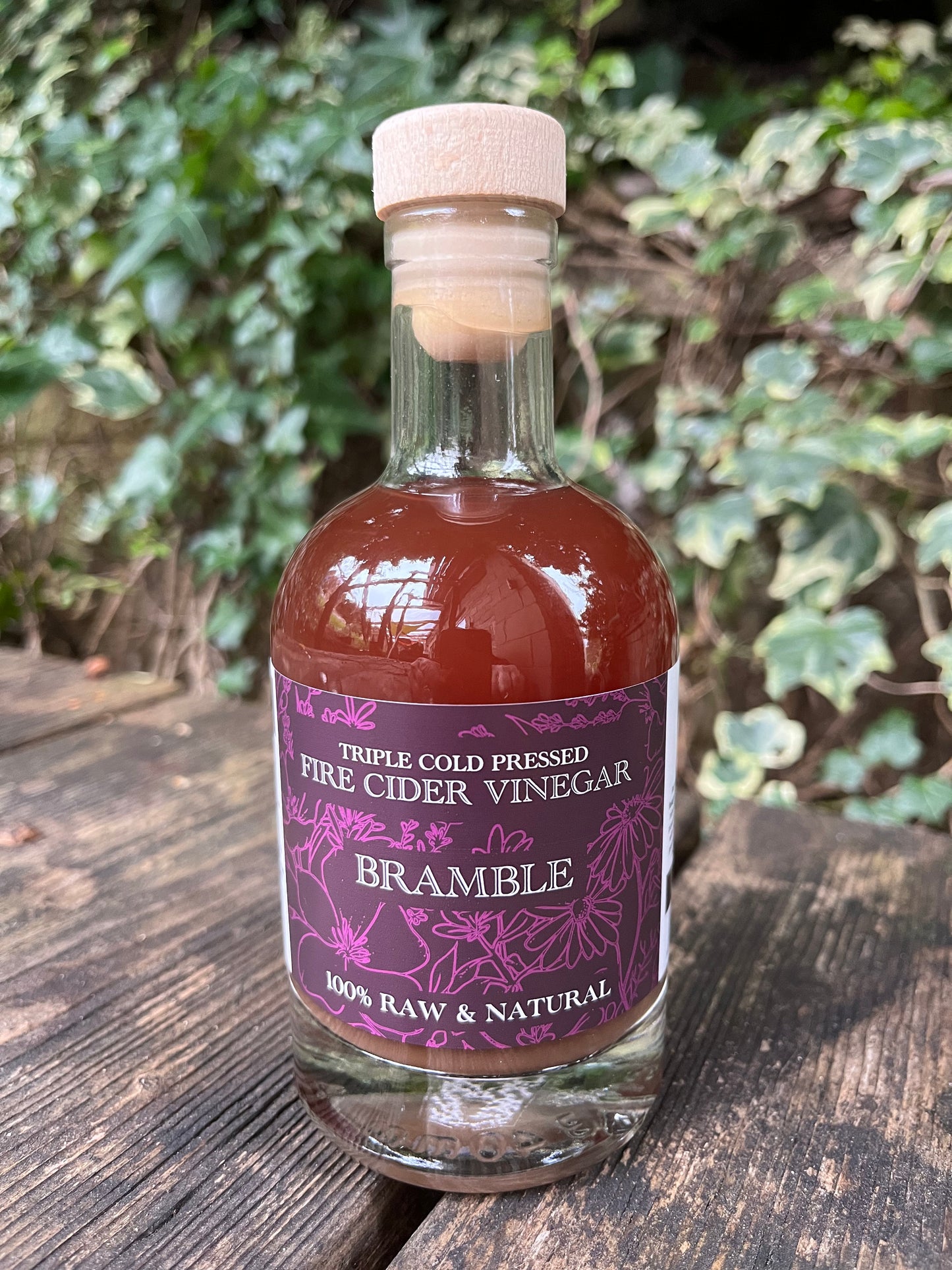 Bottle of Bramble Fire Cider Vinegar on a wooden surface with greenery in the background