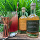 Two bottles of 'Triple Cold Pressed Fire Cider Vinegar' with a cocktail and bar tools on a wooden surface with greenery in the background.