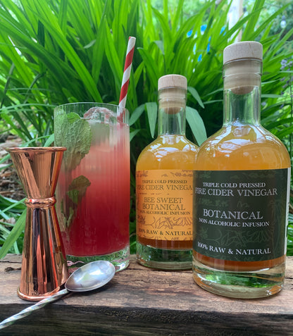 Two bottles of 'Triple Cold Pressed Fire Cider Vinegar' with a cocktail and bar tools on a wooden surface with greenery in the background.