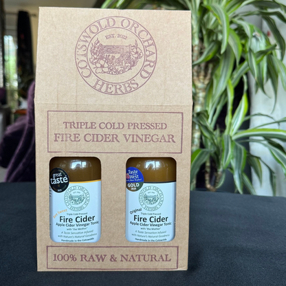 Two bottles of Fire Cider Vinegar in a branded box with a plant in the background.