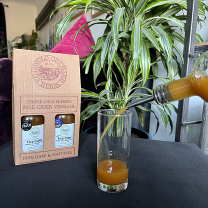 Bottle of fire cider vinegar being poured into a glass with a cardboard box labeled 'Triple Cold Pressed Fire Cider Vinegar' in the background.