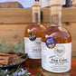 Two bottles of 'Taste of the West' Fire Cider on a wooden surface with herbs and spices.