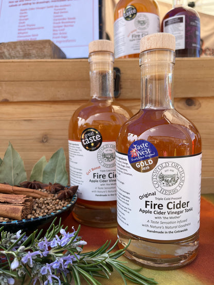 Two bottles of 'Taste of the West' Fire Cider on a wooden surface with herbs and spices.