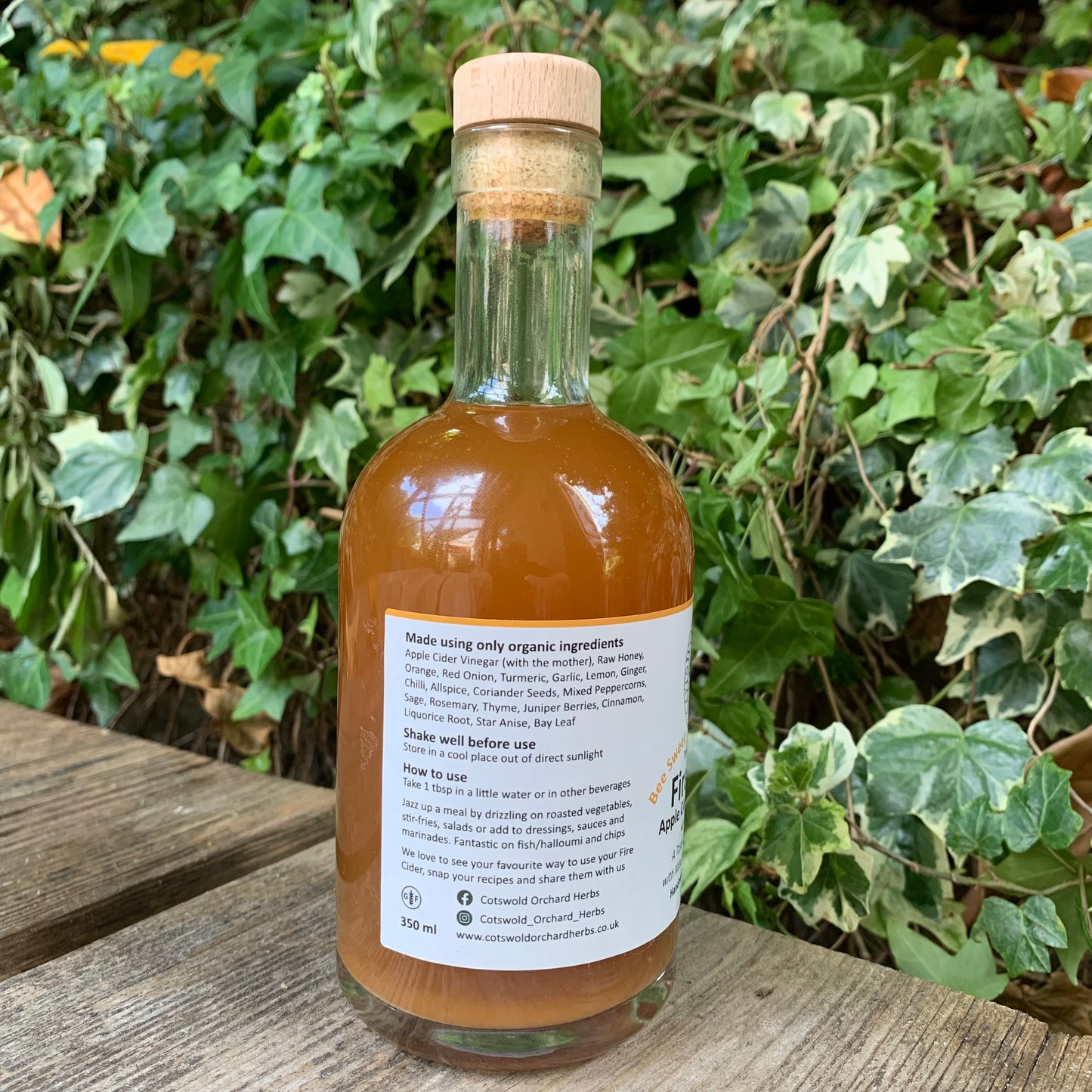 Side view of Bee Sweet Fire Cider 350ml bottle – label showing organic ingredients and usage directions, placed outdoors on a wooden bench surrounded by ivy