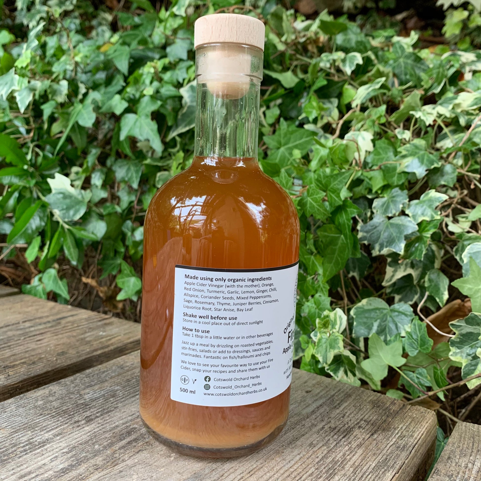 Side label of Original Fire Cider 500ml bottle – shows organic ingredients and usage instructions, with cork stopper and amber liquid, placed on wooden surface against vibrant ivy foliage